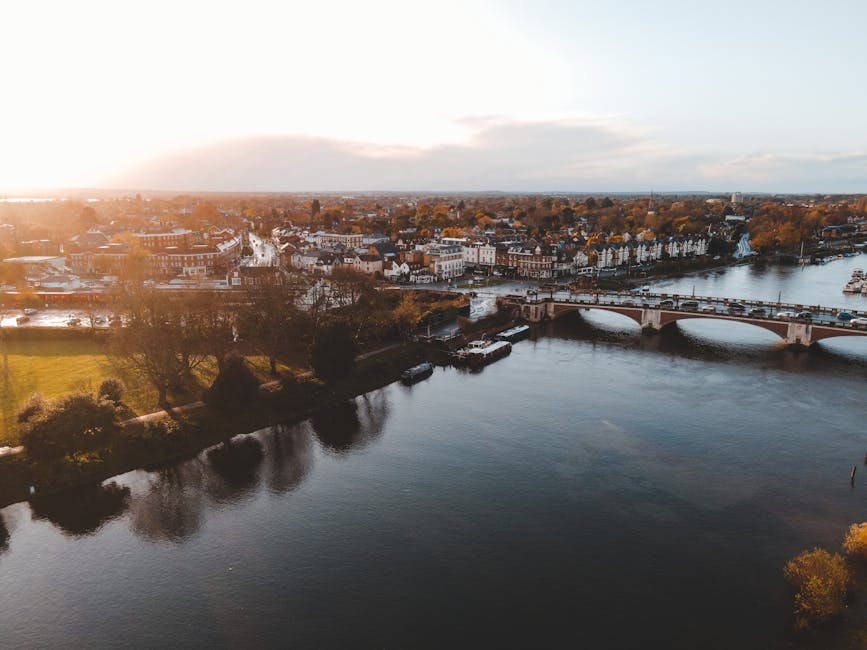 An aerial view of a river with calm water reflecting the sky, taken during sunset with warm light illuminating the scene. On one side of the river, there is a row of white residential buildings with d