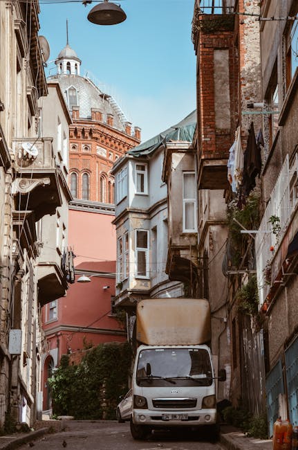 A narrow cobbled street in a residential area with closely packed houses on either side, featuring sloped tiled roofs, chimneys, and traditional street lamps. The scene appears to be outdoors during d